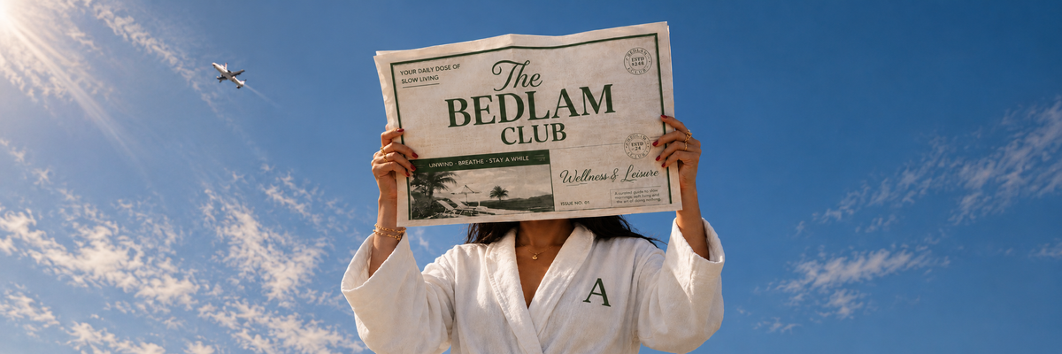 Woman in a monogrammed white BEDLAM bathrobe holding The Bedlam Club newspaper against a blue sky